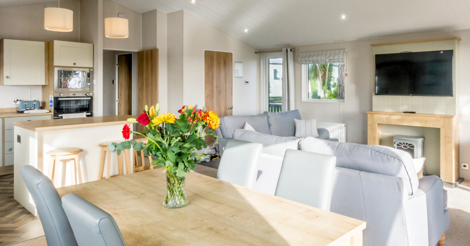 Bright open-plan living and dining area with wooden table, grey sofas and a wall-mounted TV
