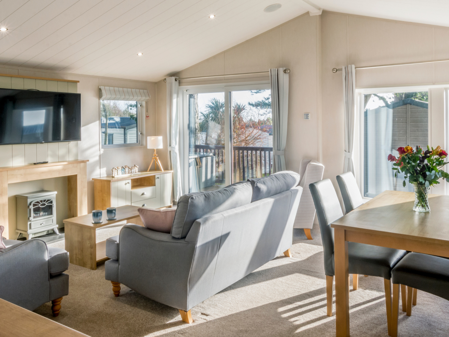 Bright, modern living and dining area with sofas, a wall-mounted television, and large glass doors leading to a deck