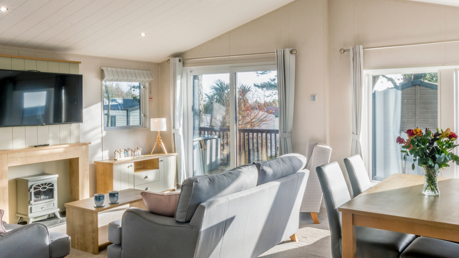 Bright, modern living and dining area with sofas, a wall-mounted television, and large glass doors leading to a deck