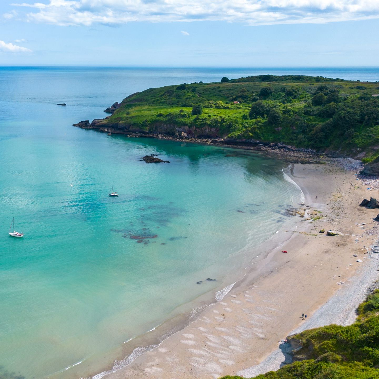 Aerial view of a turquoise bay with sandy beach, anchored boats and green headland under a blue sky