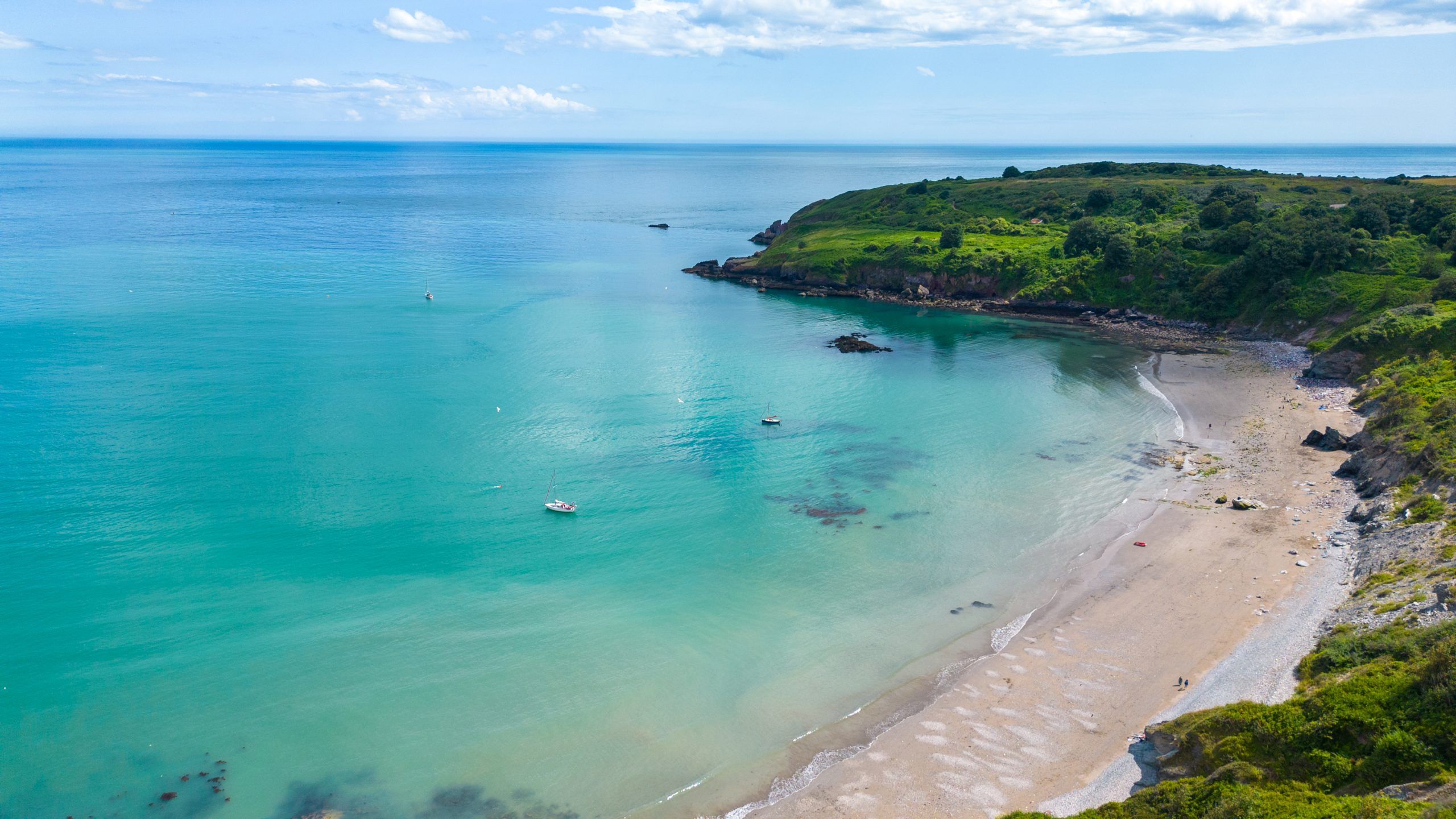 Aerial view of a turquoise bay with sandy beach, anchored boats and green headland under a blue sky