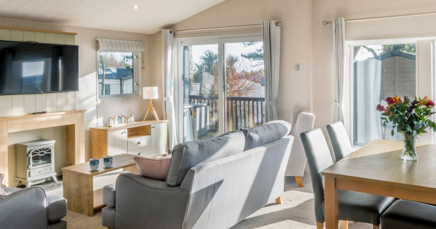 Bright open-plan living and dining area with grey sofas, wooden table, wall-mounted TV and large patio doors