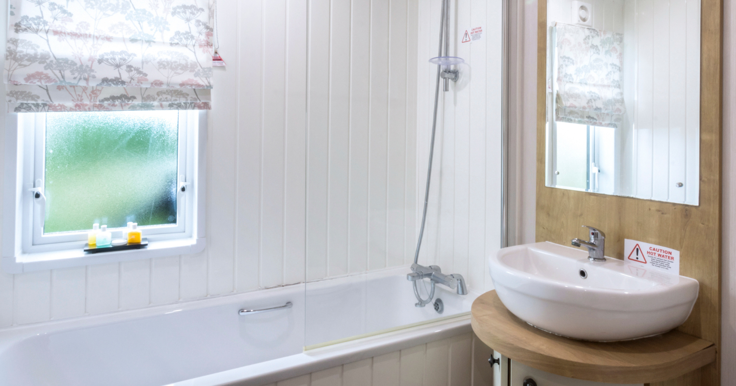 Bright white bathroom with a bath and overhead shower, window with patterned blind, and a washbasin with mirror