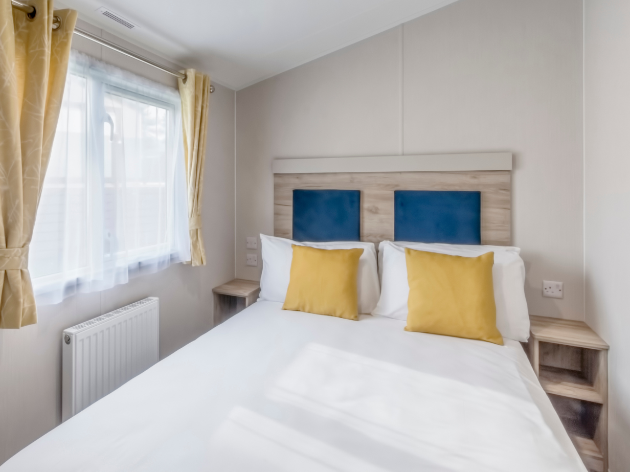 Bright double bedroom with white bedding, blue headboard panels and yellow cushions beside a window with yellow curtains.