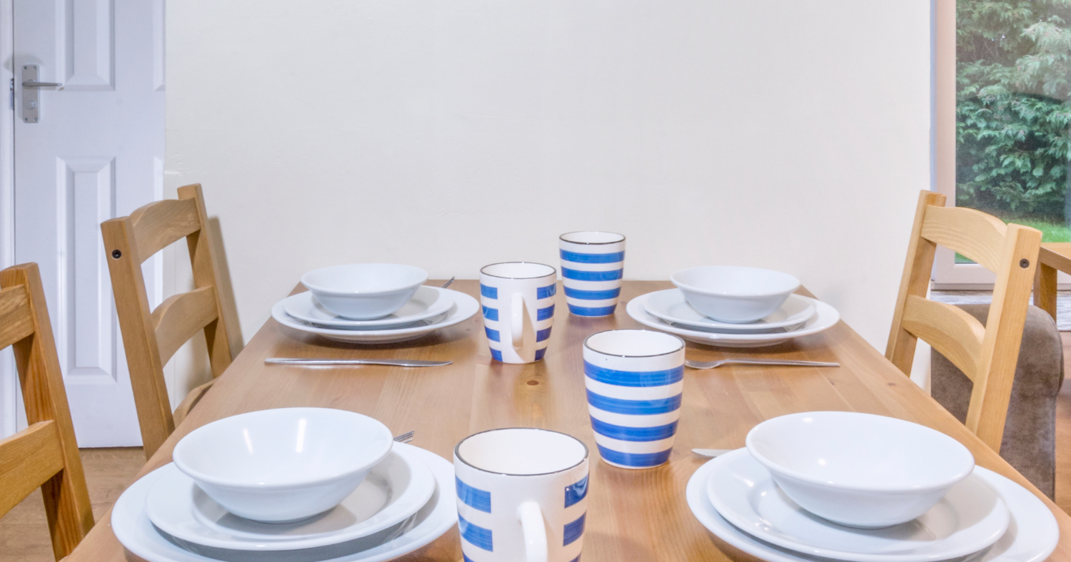 Dining table set for four with white bowls and plates, blue-striped mugs, and cutlery on a wooden table
