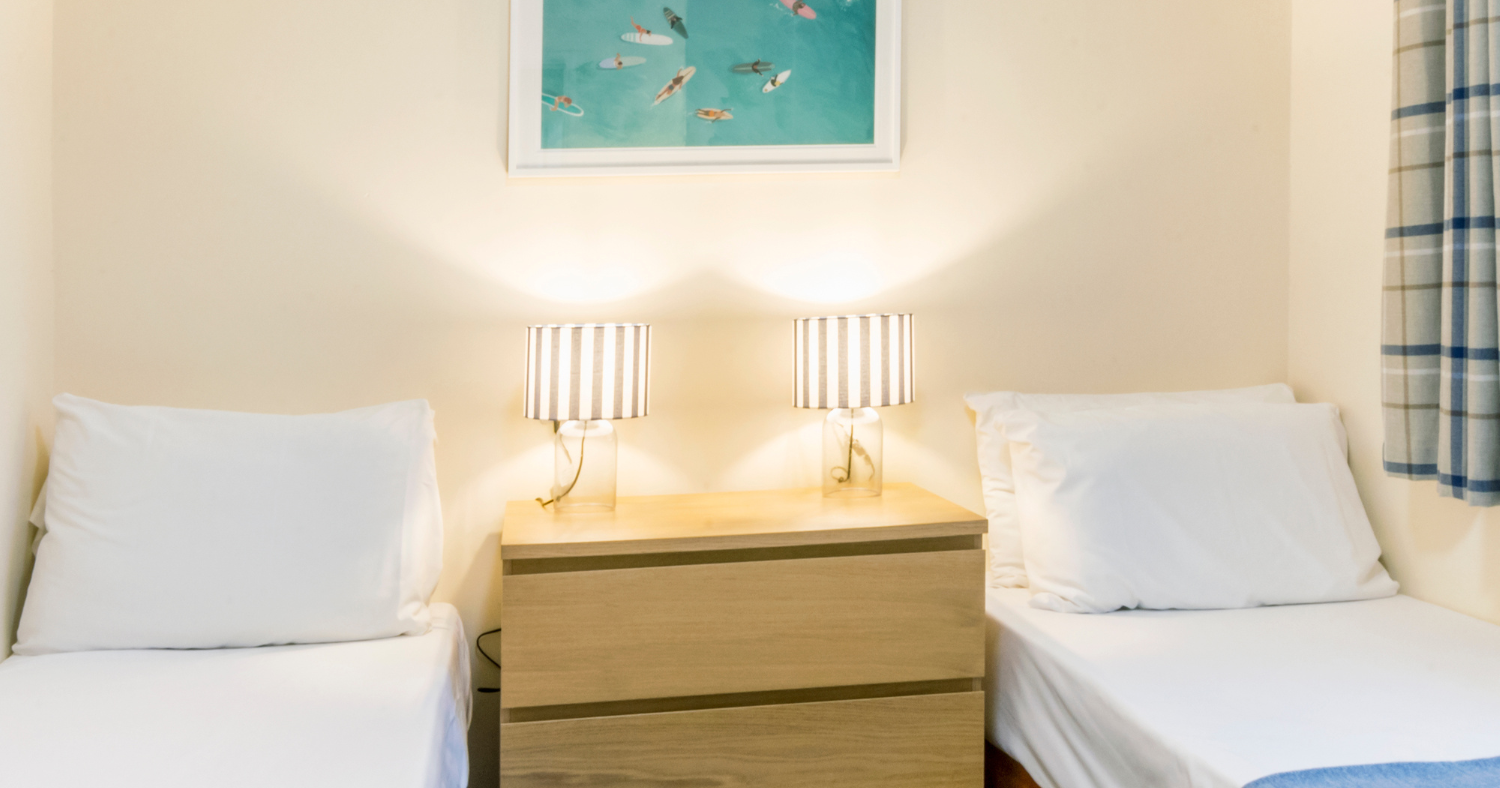 Twin beds with blue throws and bedside table with two lamps beneath framed beach artwork