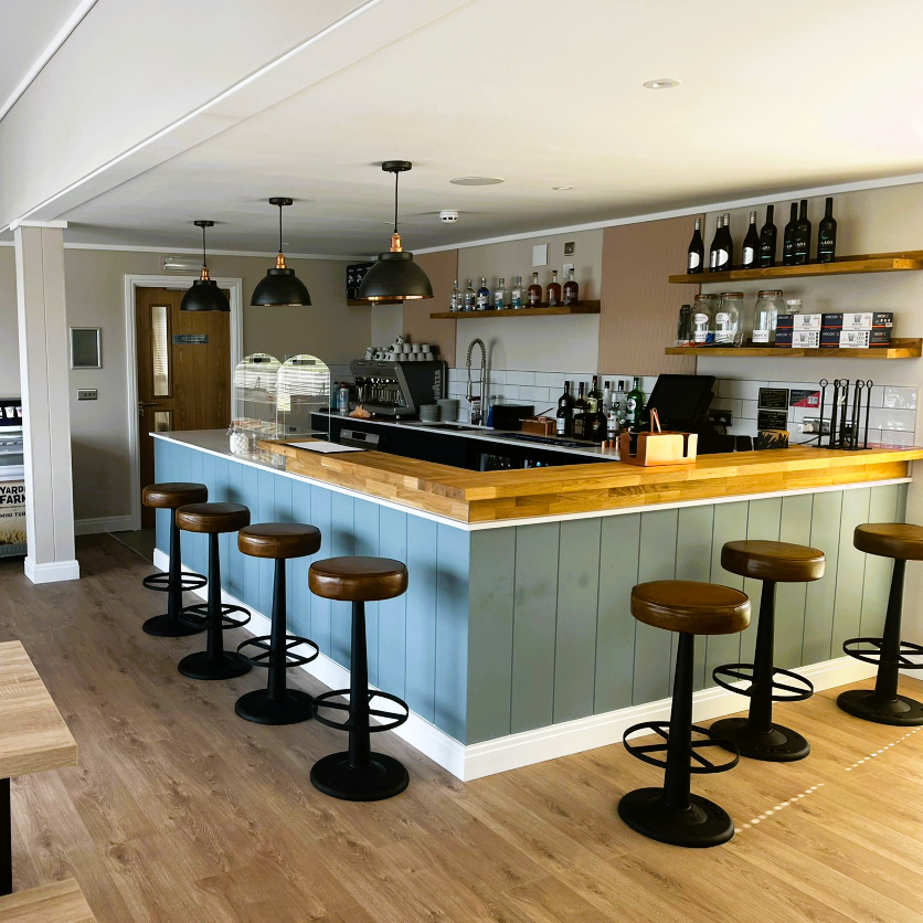 Modern bar interior with wooden countertop, pastel panelled bar front and leather bar stools under pendant lights