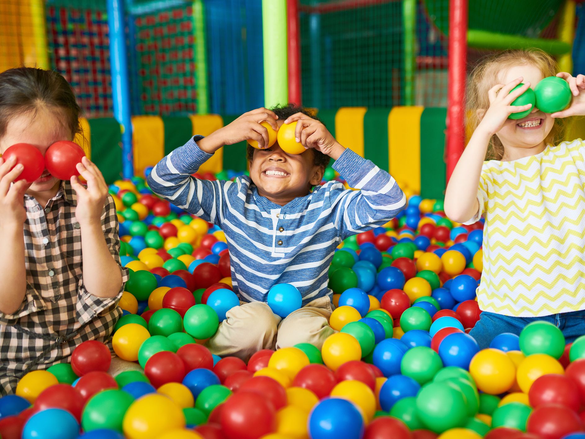 Three children playing in a colourful ball pit, holding plastic balls over their eyes