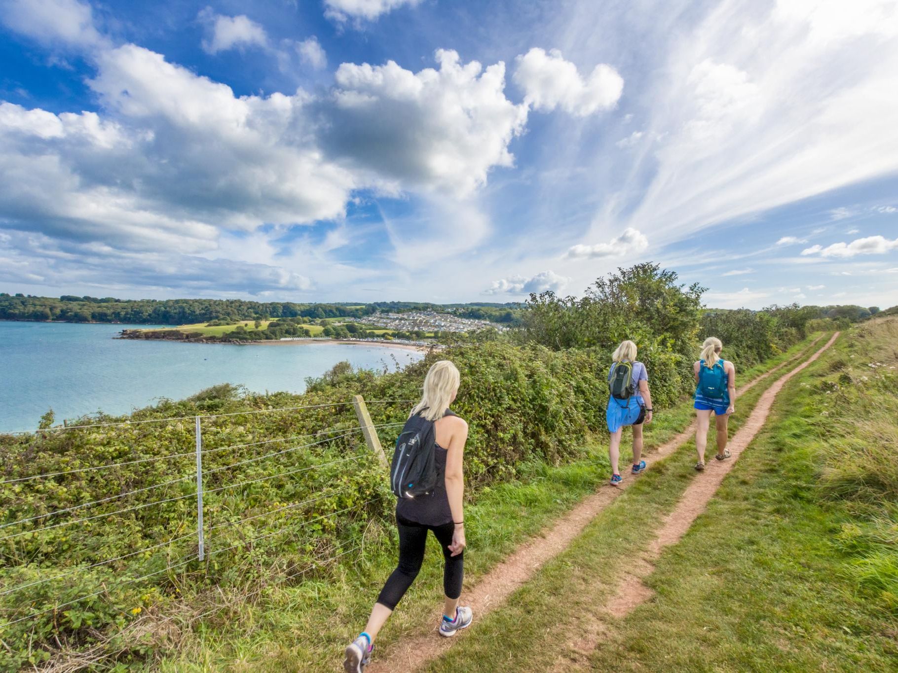 Three walkers with rucksacks on a coastal path above a blue bay under a bright, cloud-filled sky