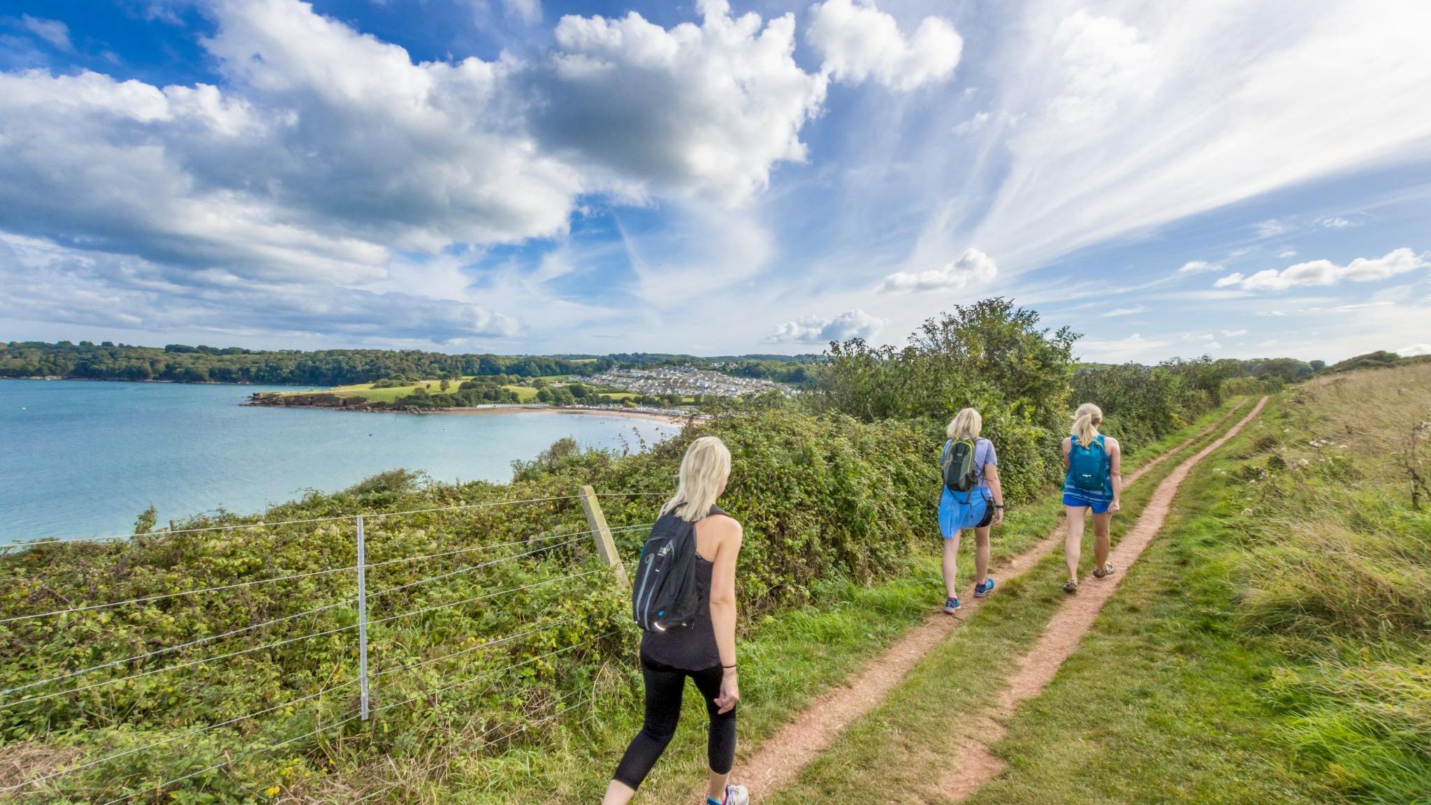 Three walkers with rucksacks on a coastal path above a blue bay under a bright, cloud-filled sky