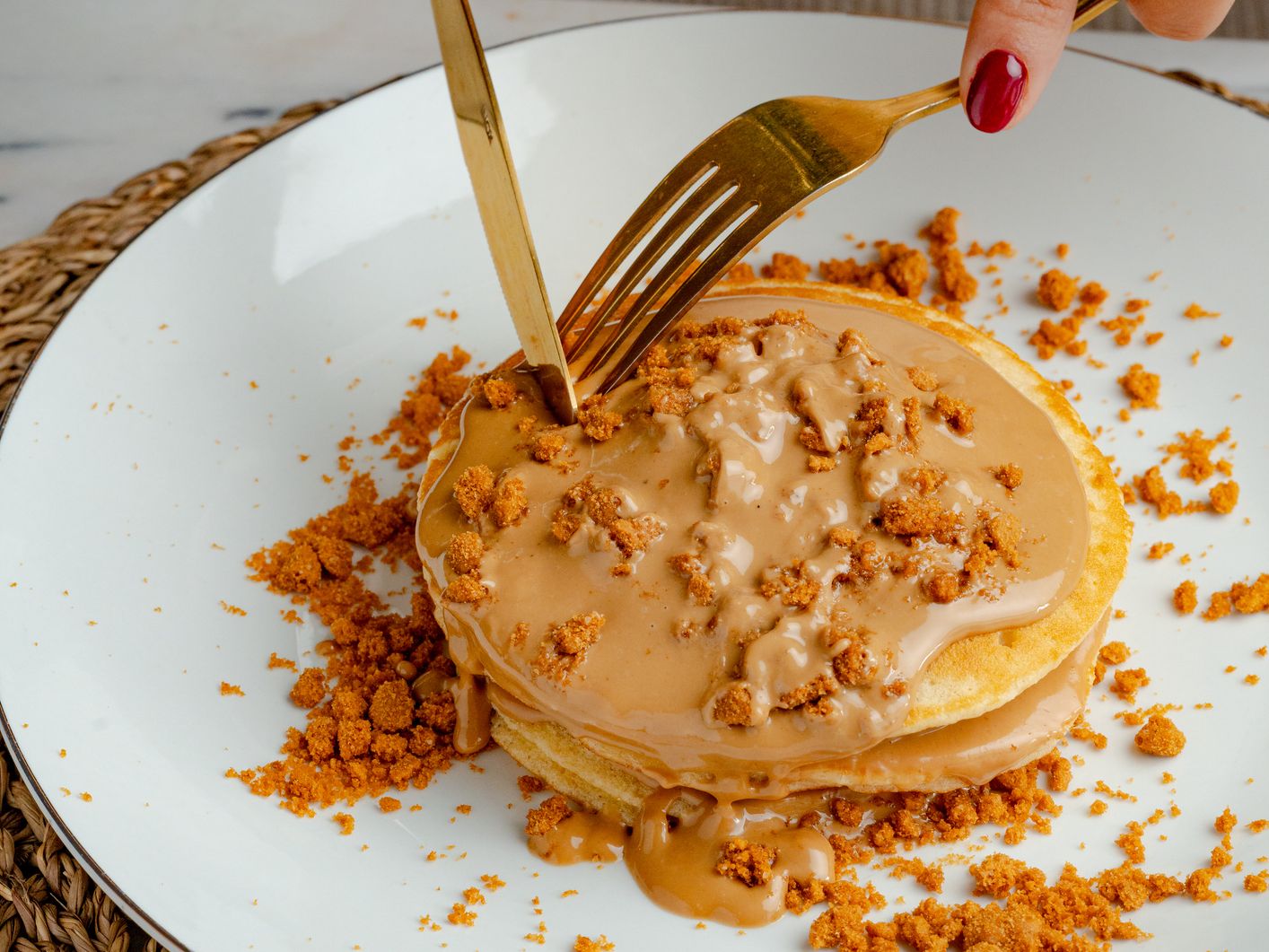 Hands cutting a stack of pancakes topped with caramel sauce and biscuit crumbs on a white plate