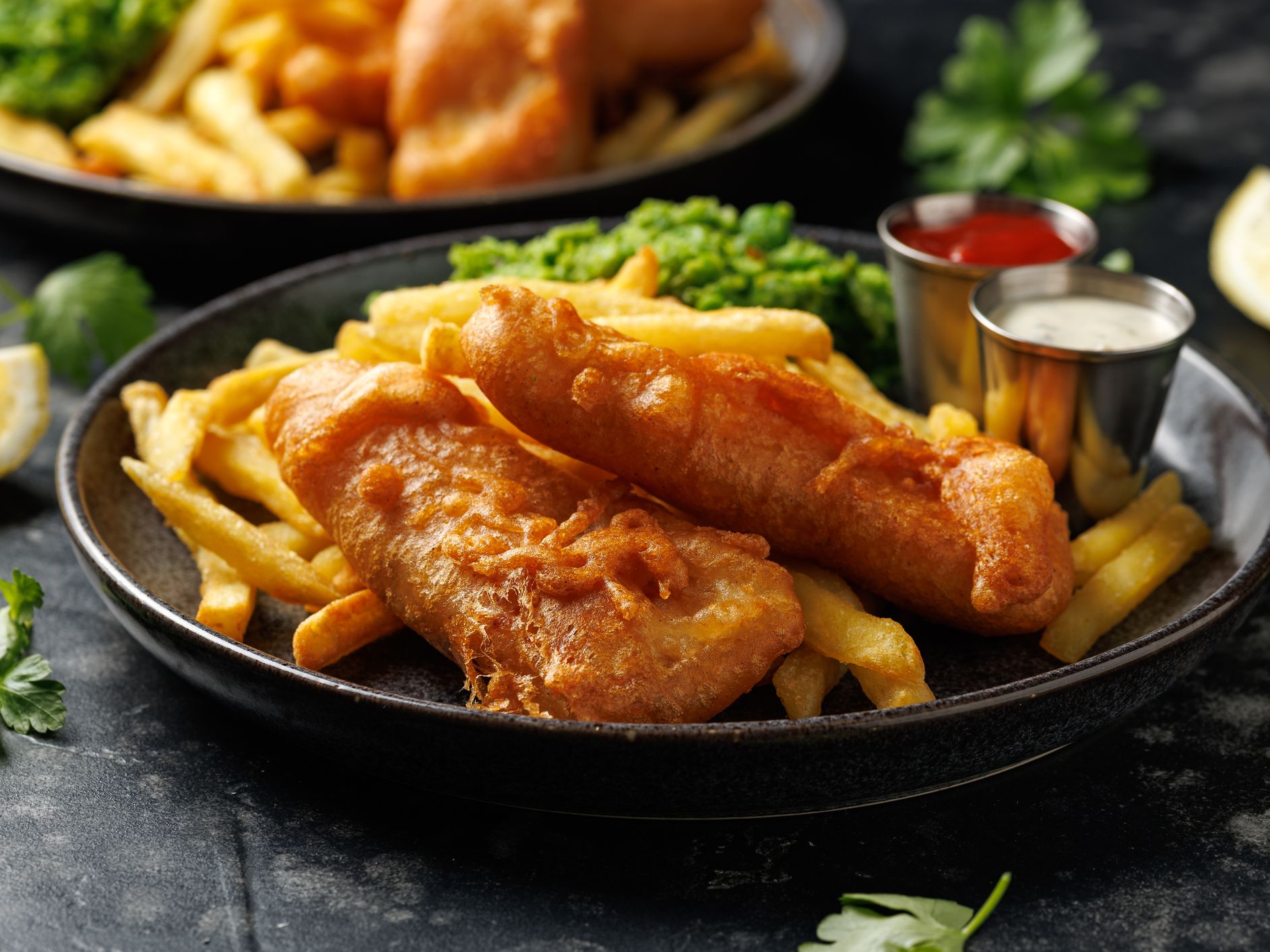 Battered fish and chips with mushy peas, tartare sauce and ketchup on a dark plate