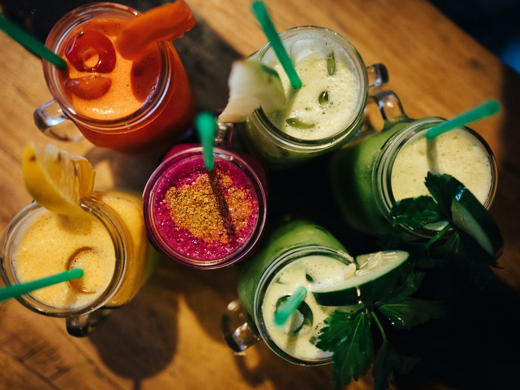 Top-down view of six colourful smoothies in glass jars with green straws on a wooden table