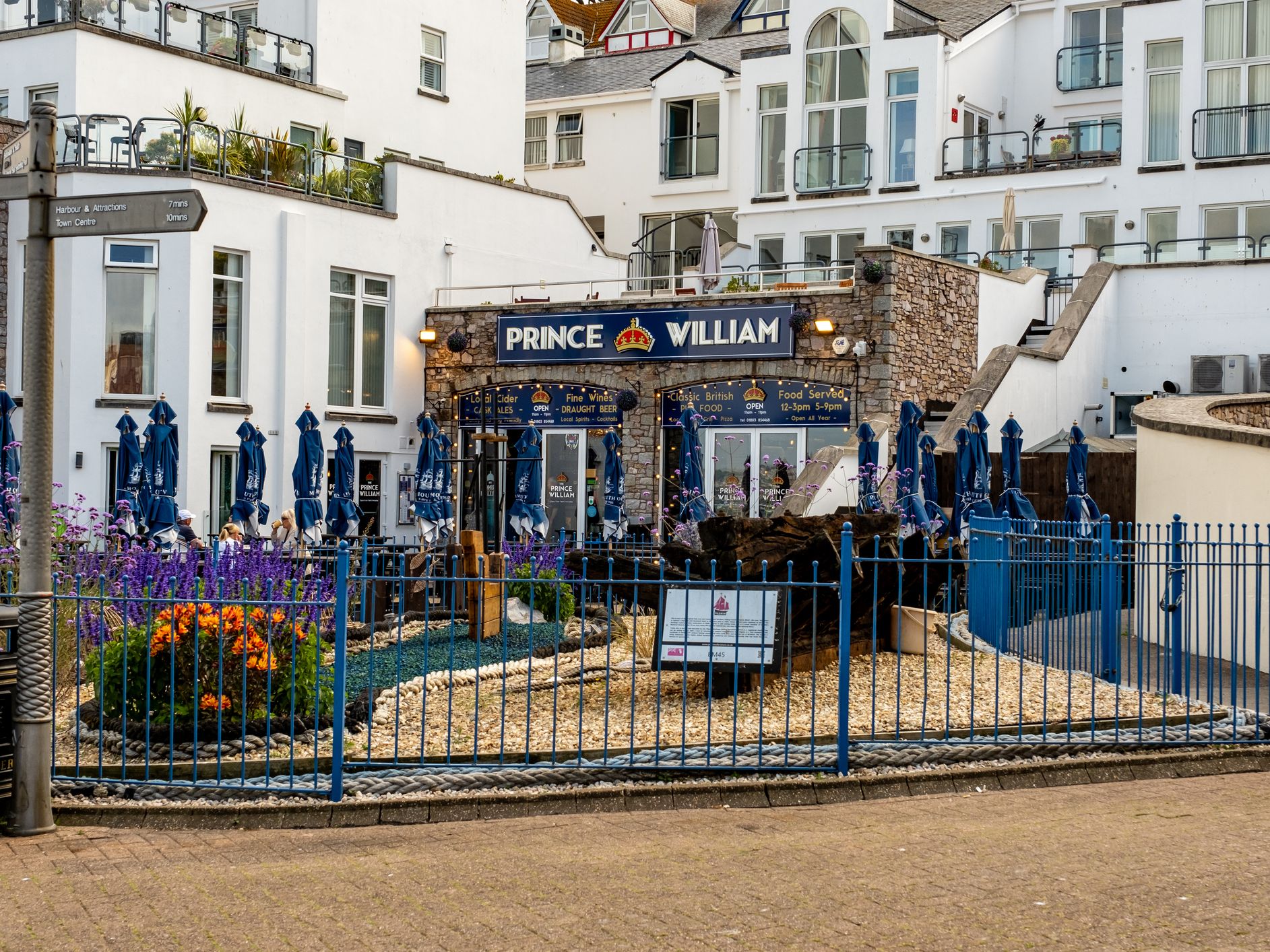 Exterior of the Prince William pub with blue umbrellas and a fenced flower bed in front