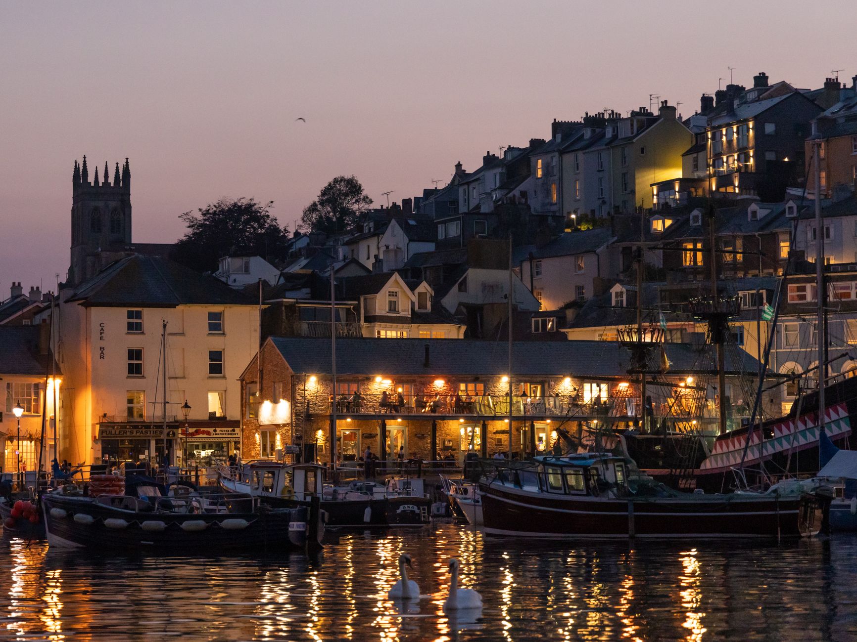 Harbour at dusk with moored boats, lit waterside buildings and hillside houses under a purple sky