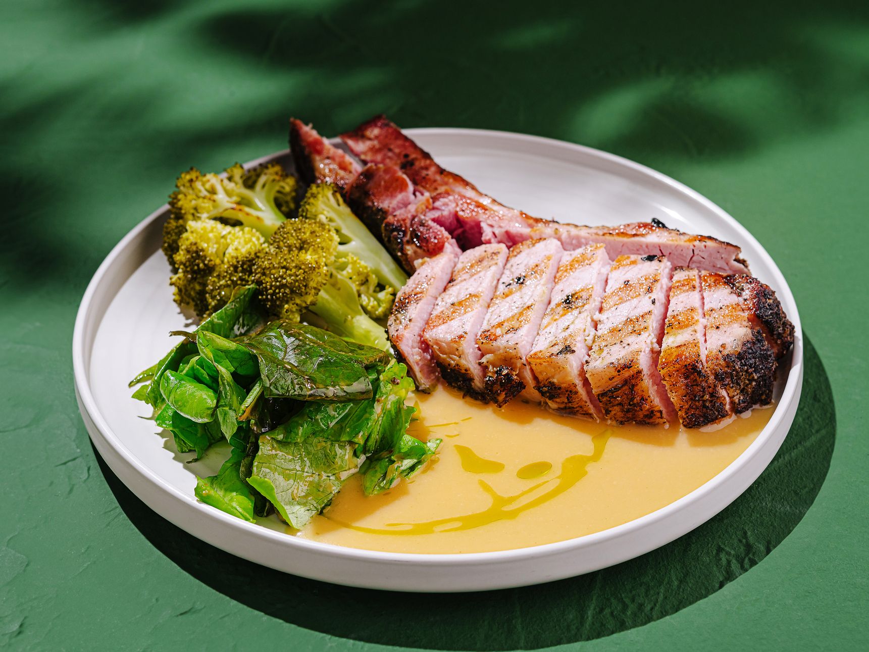 Sliced grilled pork with broccoli and leafy greens on a white plate with sauce