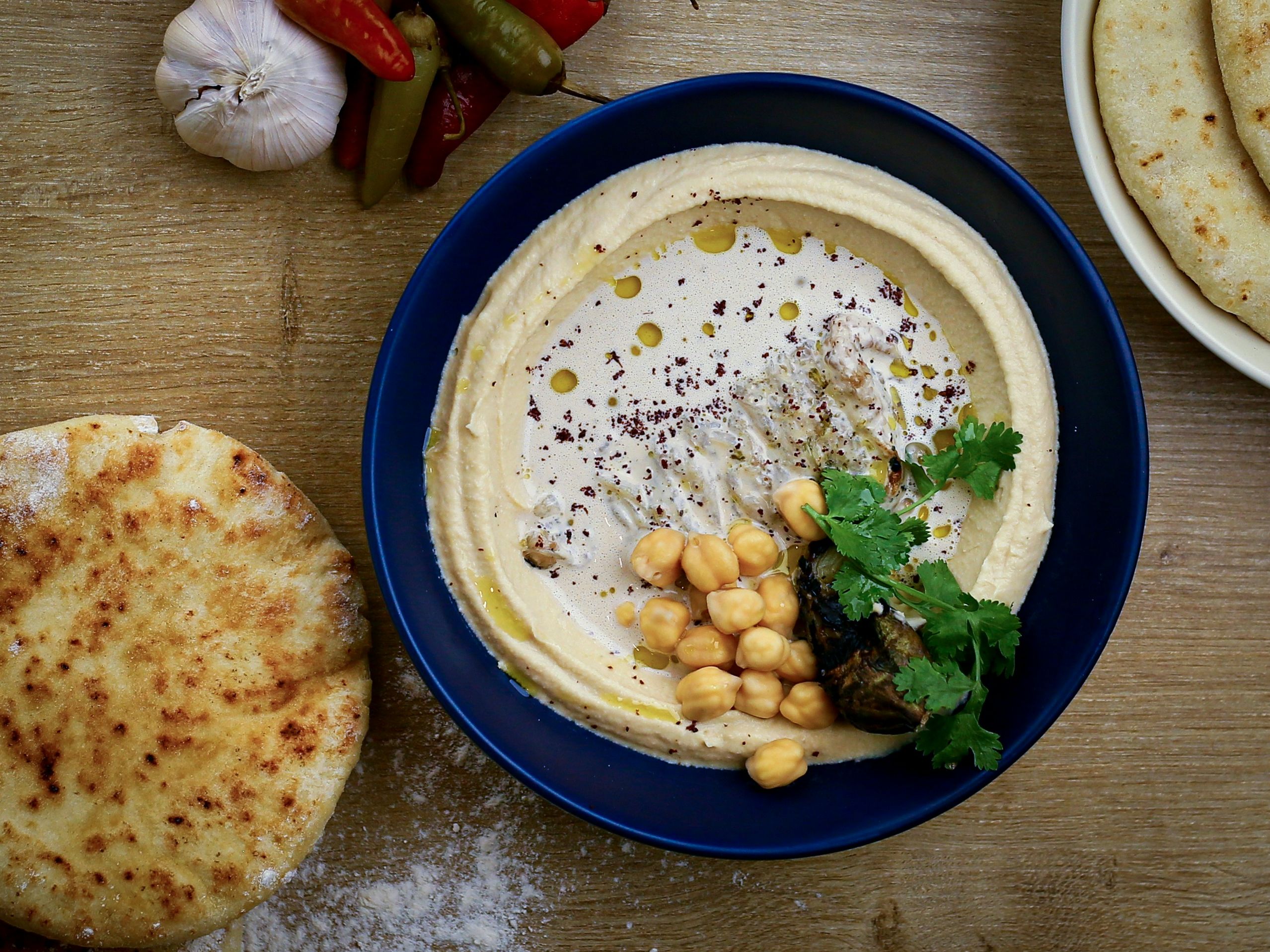 Bowl of creamy hummus topped with chickpeas, olive oil, spices and fresh coriander, served with flatbreads on a wooden table.