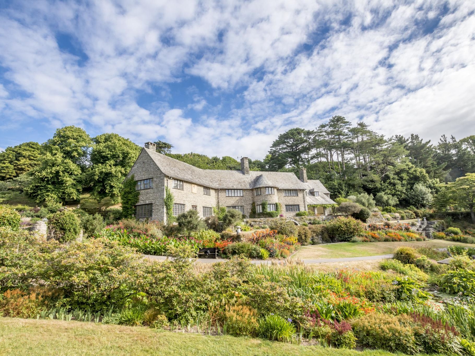 Large stone country house set above colourful landscaped gardens under a bright blue sky with scattered clouds
