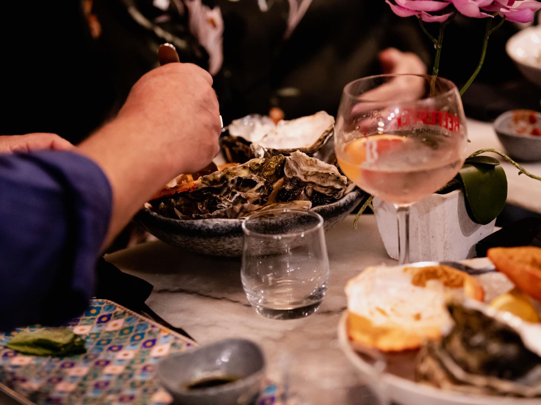 Hands serving oysters from a bowl on a marble table beside a glass of rosé and a small vase of pink flowers.