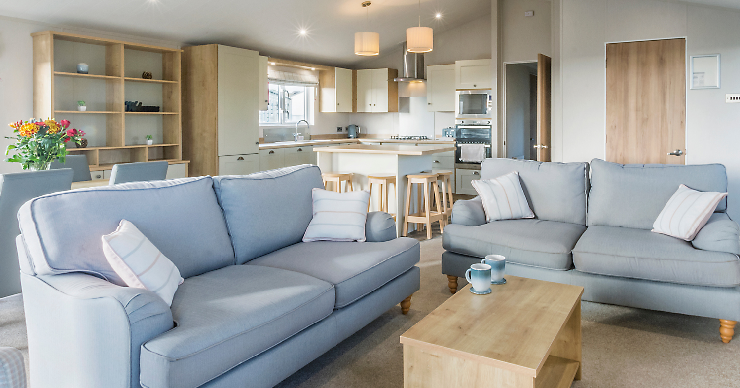 Open-plan lodge lounge with two blue sofas, wooden coffee table and modern kitchen with breakfast bar