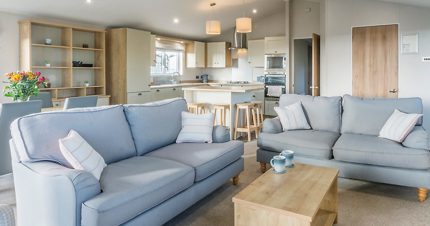 Open-plan lodge lounge with two blue sofas, wooden coffee table and modern kitchen with breakfast bar