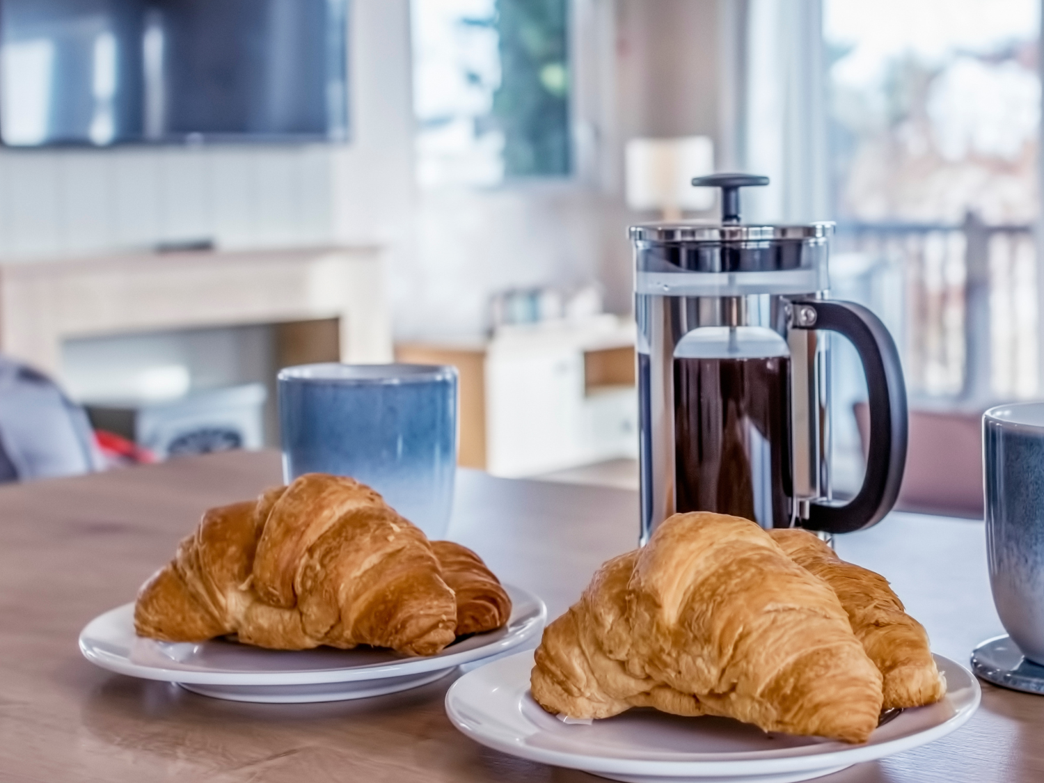 Croissants on plates with a French press coffee pot and mugs on a table in a bright lodge living room.