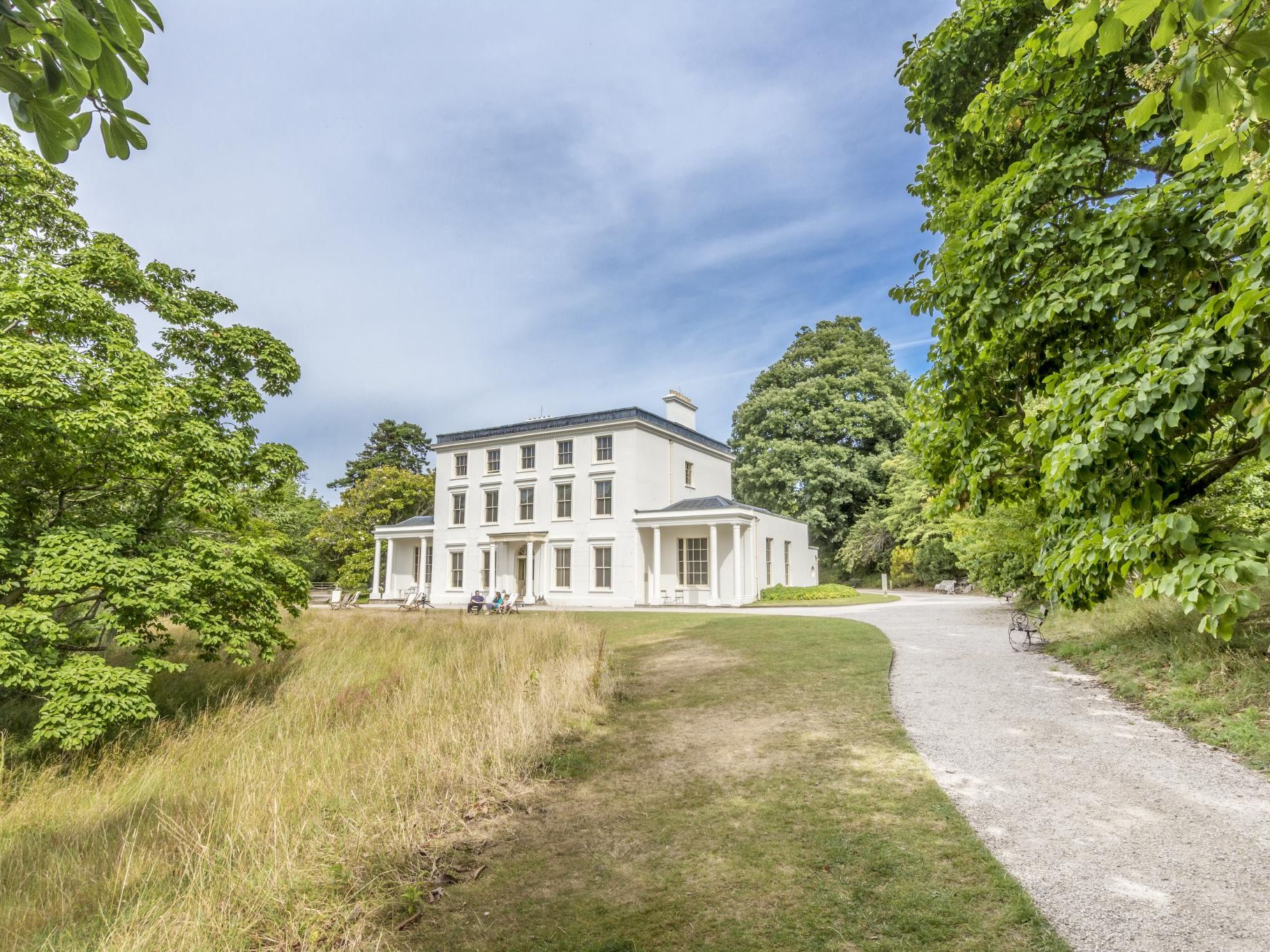 Large white country house with tall windows, surrounded by trees and a curved gravel path