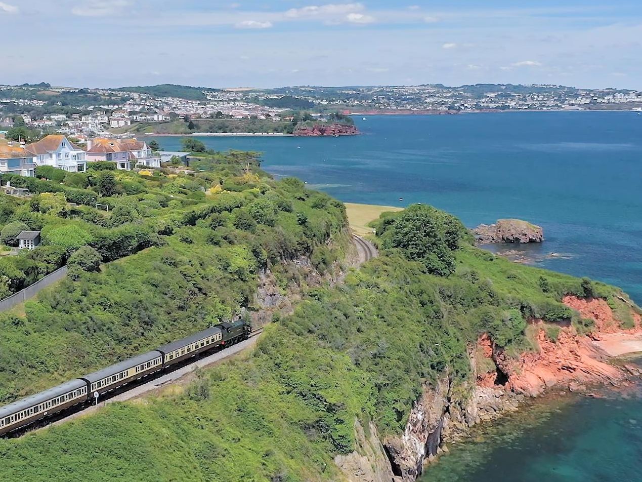 Train travelling along a coastal cliff railway beside blue sea and hillside houses