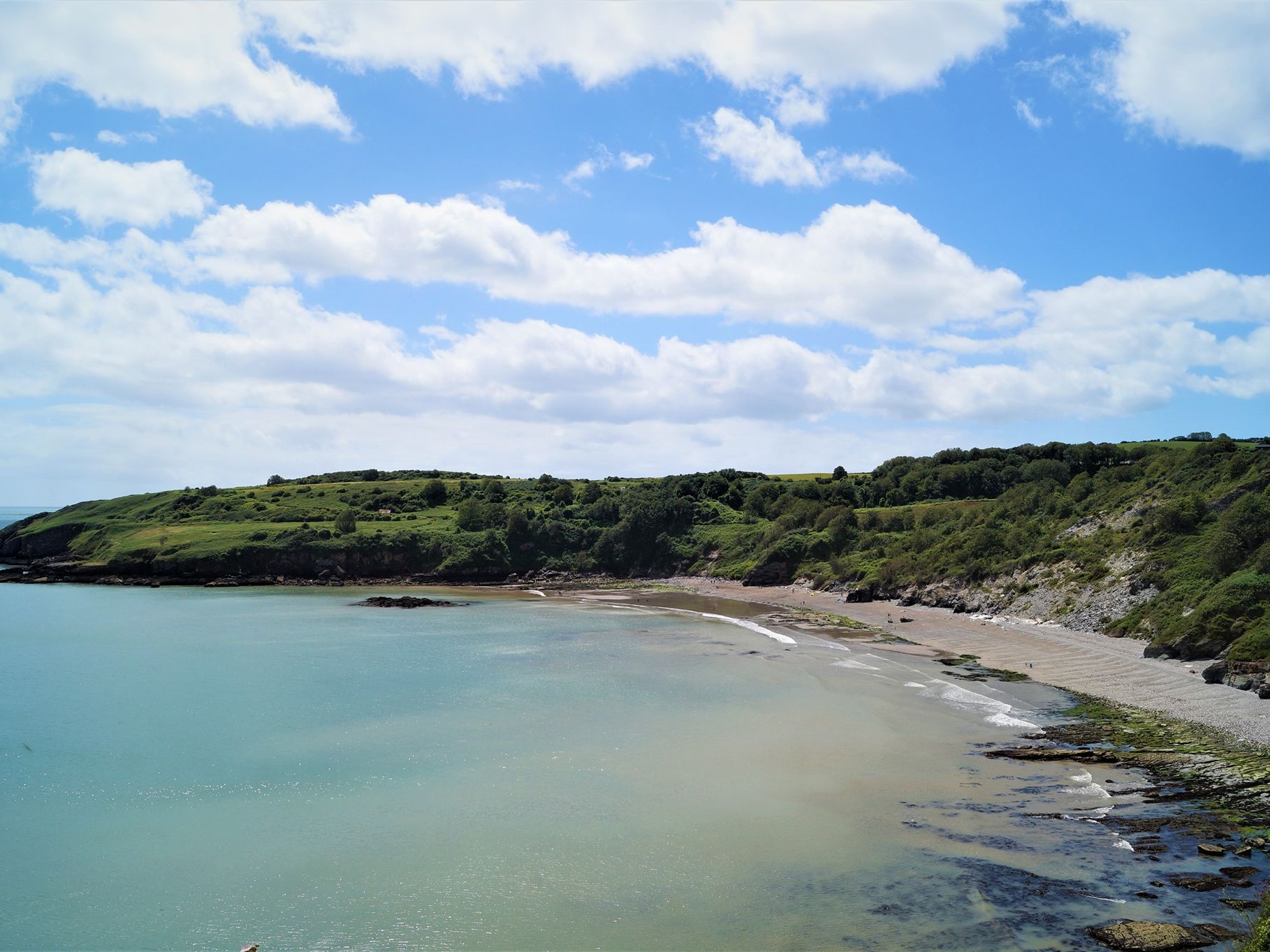 Coastal bay with sandy beach, rocky shoreline and green hills beneath a blue sky with scattered clouds
