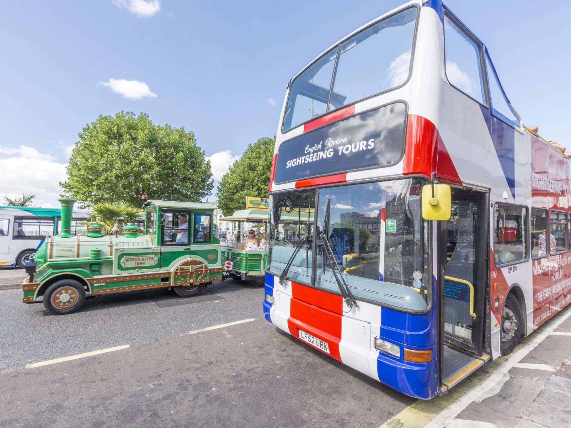 Open-top English Riviera sightseeing tour bus parked beside a green tourist road train on a sunny day