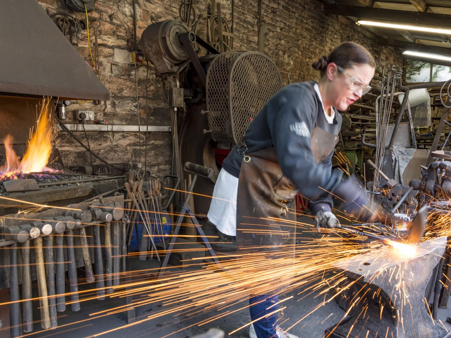 Blacksmith at Cockington Court and Craft Centre shaping hot metal on an anvil as sparks fly, with a lit forge in the background.