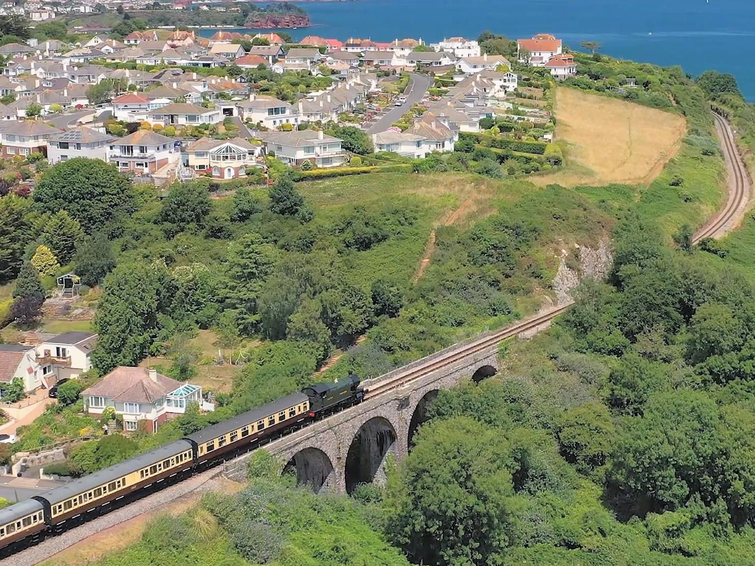 Aerial view of a steam train crossing a stone viaduct on the Dartmouth Steam Railway beside the coast