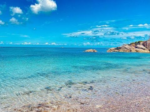 Clear turquoise sea lapping a pebbled beach beside a rocky breakwater under a bright blue sky