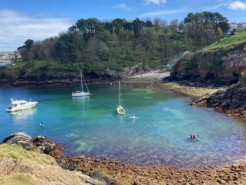 Boats anchored in the clear turquoise waters of Churston Cove, with rocky shoreline and wooded hillside in the background.