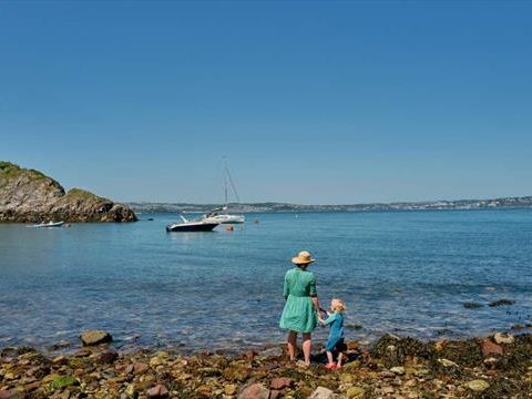 Adult and child on a rocky shoreline at Fishcombe Cove, looking out over calm blue sea with a moored sailboat