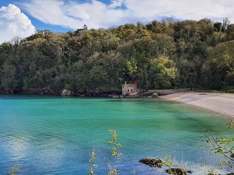 Turquoise sea in a sheltered cove with a sandy beach, small stone hut and wooded hillside under a partly cloudy sky