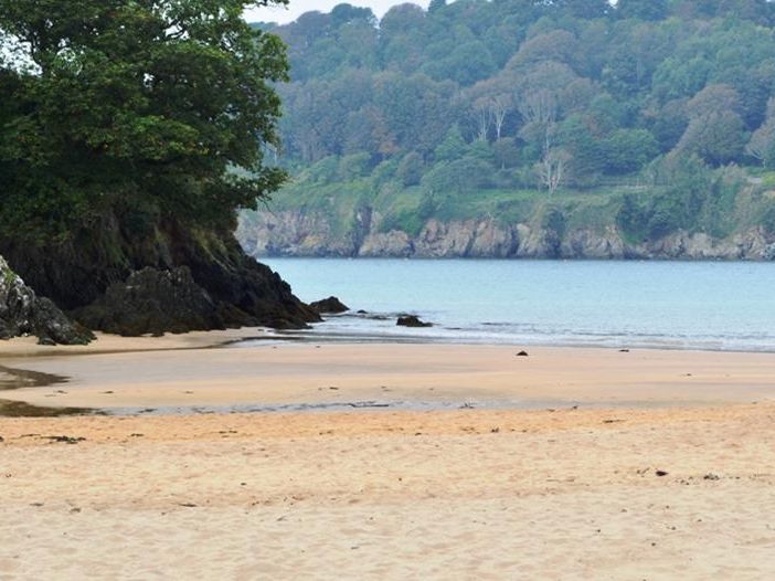 Sandy beach at Mill Bay Cove with calm blue water, rocky outcrops and wooded hillside beyond