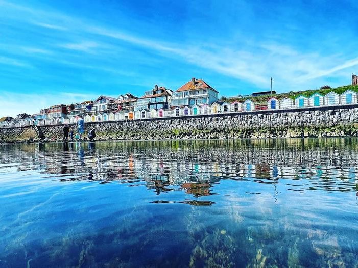 Calm low-tide sea reflecting colourful beach huts and houses along the sea wall at Preston Sands under a bright blue sky