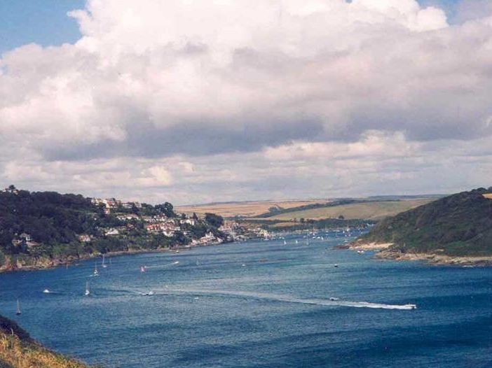 Wide view of a coastal estuary with sailboats and a speedboat wake, wooded hills and a village under a cloudy sky