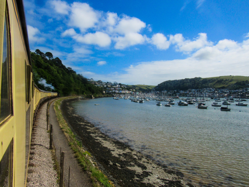 A train travels along the coast, overlooking a bay filled with boats under a blue sky.