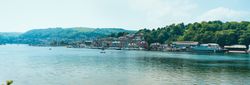 A scenic view of a riverside town with colorful buildings along the water, boats docked by the shore, and green hills in the background under a clear, bright sky.