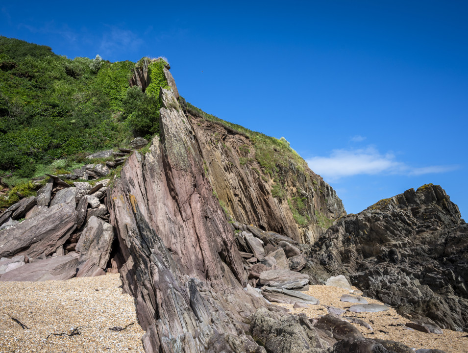 Rocky coastal cliffs covered in greenery rise above a small sandy beach under a clear blue sky, with large jagged rock formations scattered along the shoreline.