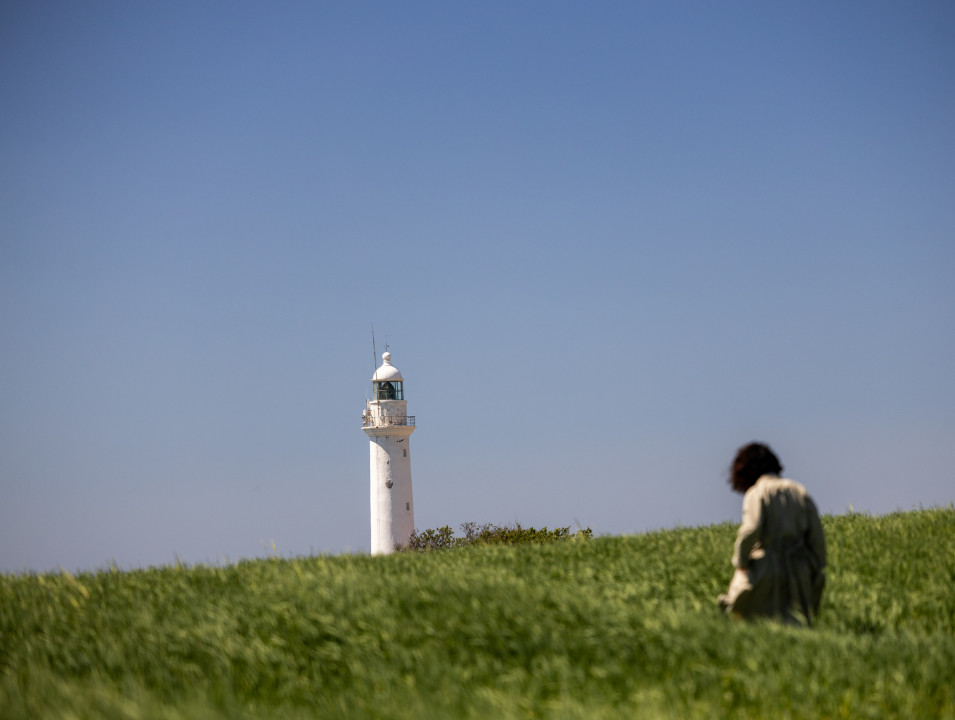 A white lighthouse stands tall against a clear blue sky, partially obscured by a grassy field, with a person in a coat walking through the grass in the foreground.