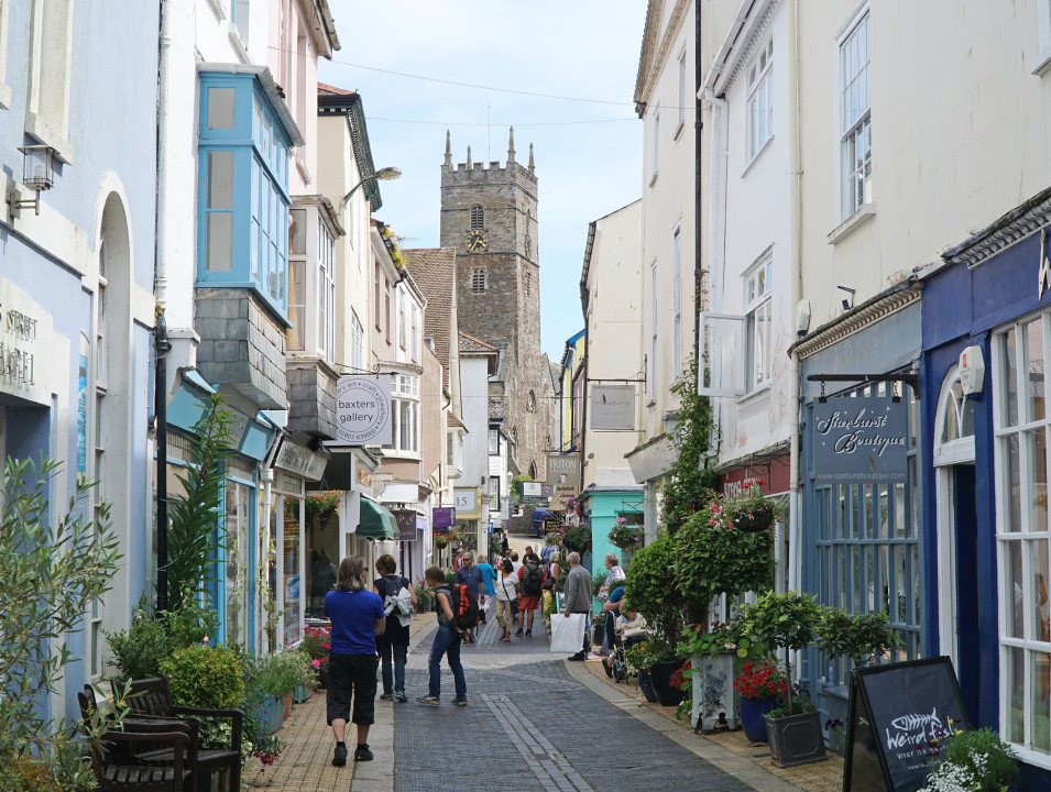 A charming street scene in a historic town, lined with colorful shops and signs. People stroll along the narrow cobbled path, with a stone church tower visible in the background.