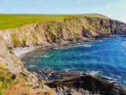 A rugged coastal landscape featuring steep cliffs and clear turquoise waters. The rocky shoreline meets grassy hills, with waves gently lapping against the shore under a bright sky.