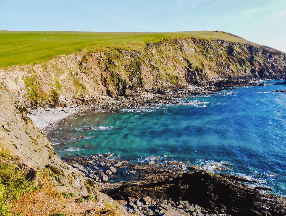 A rugged coastal landscape featuring steep cliffs and clear turquoise waters. The rocky shoreline meets grassy hills, with waves gently lapping against the shore under a bright sky.