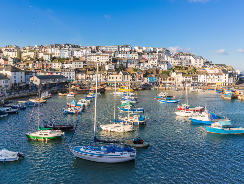 Brixham harbor filled with colorful boats, surrounded by pastel-colored buildings on a hillside. The bright blue sky reflects off the calm water, creating a vibrant seaside scene.