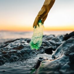 A gloved hand picks up a green plastic bottle from a rocky shoreline at sunset, with gentle ocean waves and an orange sky in the background.