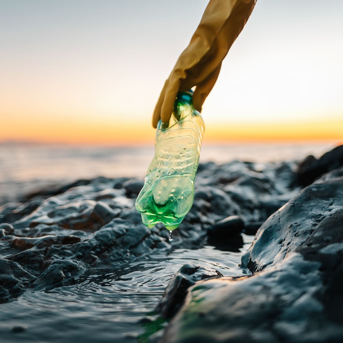 A gloved hand picks up a green plastic bottle from a rocky shoreline at sunset, with gentle ocean waves and an orange sky in the background.
