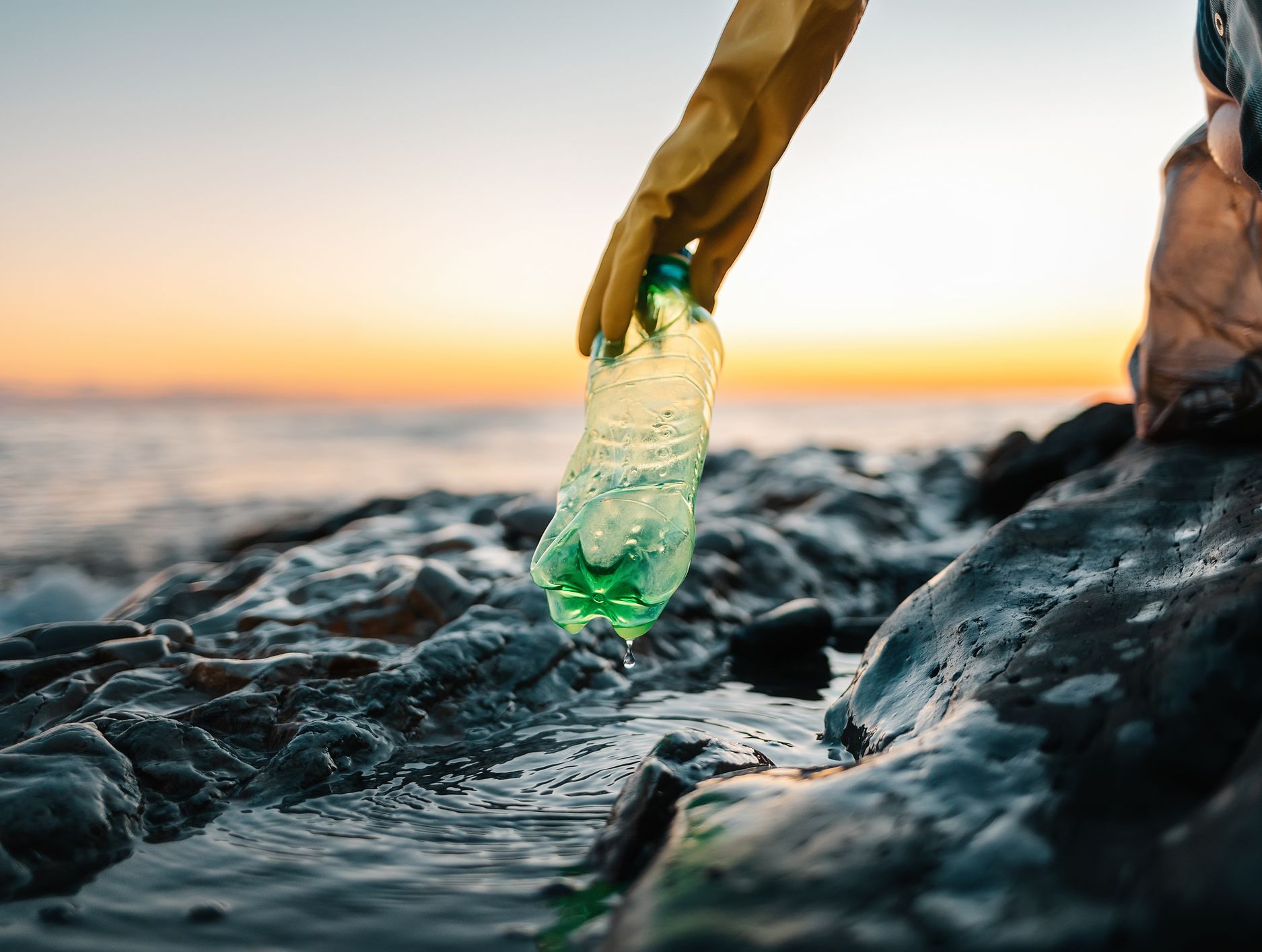 A gloved hand picks up a green plastic bottle from a rocky shoreline at sunset, with gentle ocean waves and an orange sky in the background.